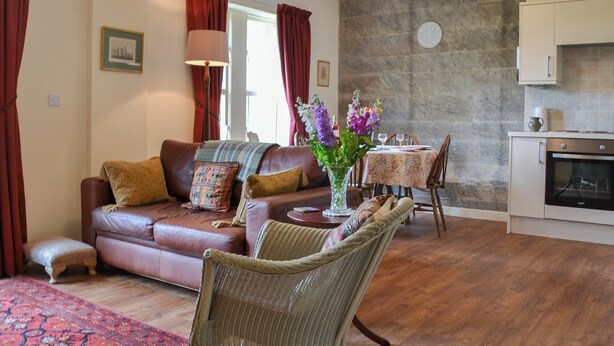 A cozy living room with a sofa, a wicker chair, a dining table with chairs, and a kitchen area with cabinets and a stove. - 2 bedroom accommodation in Brancepeth, near Durham (Brancepeth, near Durham)
