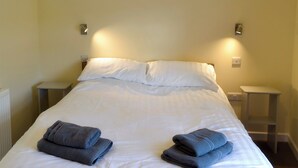 Ein ordentlich bezogenes Bett mit weißen Leinen und zwei gefalteten Handtüchern. Es befinden sich jeweils ein Nachttisch an beiden Seiten des Bettes.
