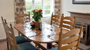 A dining room with a wooden table set for six, surrounded by wooden chairs with blue cushions. A vase with flowers and a bottle of wine are on the table.