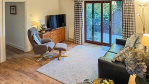 Una sala de estar con sofá, sillón y reposapiés, un televisor sobre un mueble de madera, y vistas a un espacio exterior a través de un ventanal.