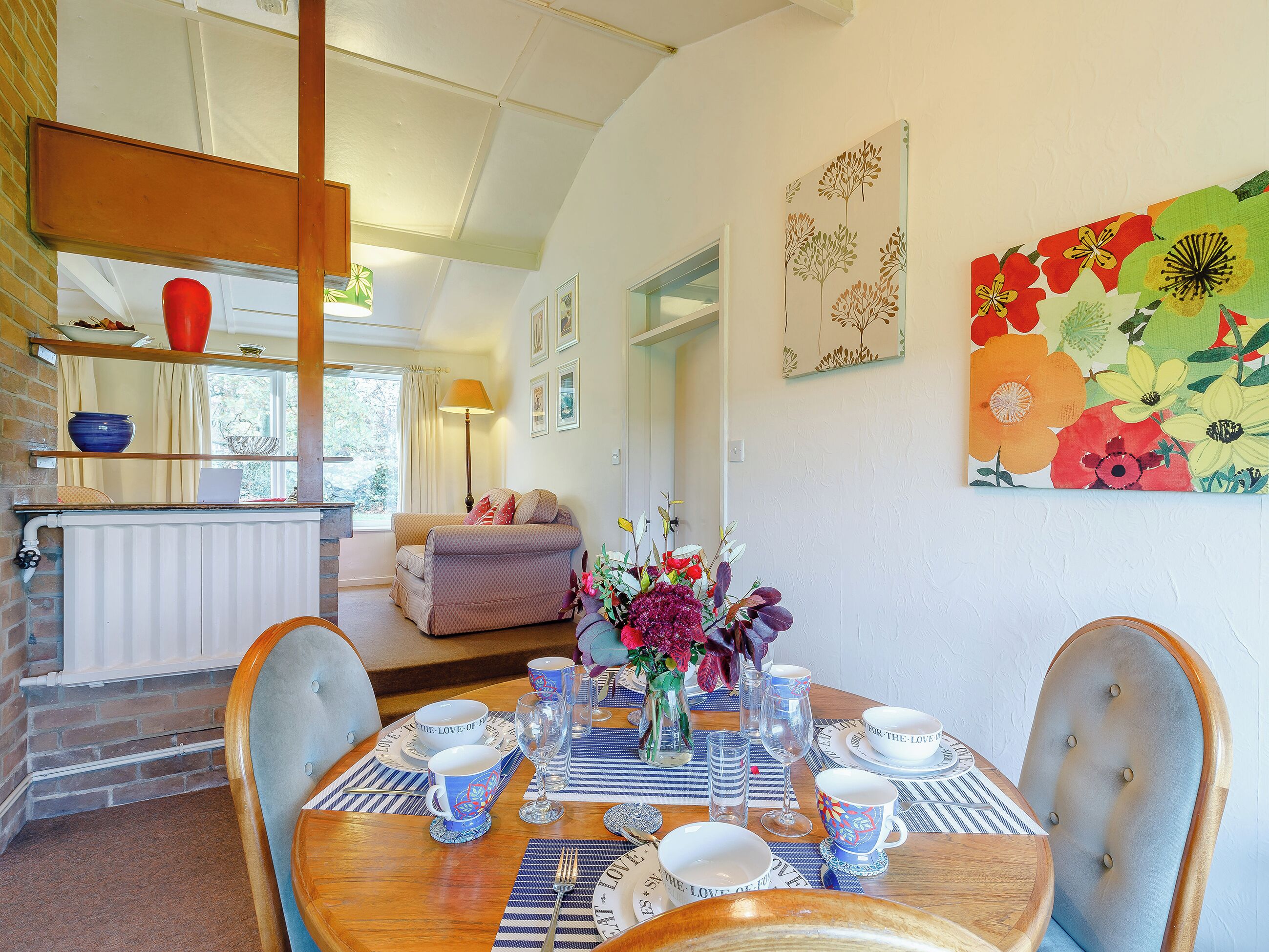 A dining area with a wooden table set for a meal, a sofa in the background, and a window with curtains.