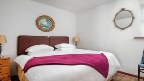 Una cama bien hecha con sábanas blancas y una manta morada, una mesita de noche de madera con una lámpara, un aparador de madera, una alfombra estampada y un cuadro en la pared.