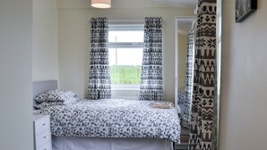 Une chambre à coucher avec un lit, une table de chevet, une fenêtre avec des rideaux à motifs et une vue sur la verdure à l’extérieur.