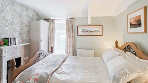 A neatly arranged bedroom with a bed, a bedside table, a radiator, a window with curtains, and a wall-mounted picture. - 3 bedroom accommodation in Aberdaron, Lleyn Peninsula (Aberdaron, Lleyn Peninsula)