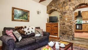 A living room with a dark patterned sofa, a painting, a stone archway, a television, and a kitchen visible through the archway.