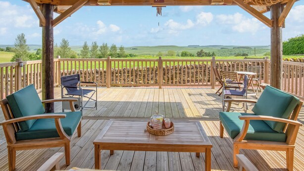 A wooden deck with outdoor seating, a table, and a stone wall in the background.