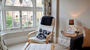 A cozy room with a large window, a chair with a cushion, a small table with a lamp, and a sofa.