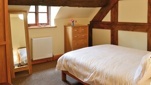Une chambre avec un plafond en pente, des poutres en bois, un lit, une commode, un miroir et une fenêtre.