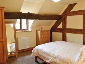 A bedroom with a sloped ceiling, wooden beams, a bed, a dresser, a mirror, and a window.