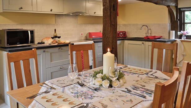 Un coin repas avec une table dressée pour quatre personnes, un centre de table composé d’une bougie et une cuisine en arrière-plan.