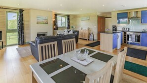 A spacious living area with a sofa, a dining table with chairs, a kitchen with modern appliances, and a view of the outdoors through a large window. - 2 bedroom accommodation in Holsworthy, near Bude (Holsworthy, near Bude)