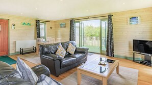 A cozy living room with a leather sofa, a coffee table, and a TV. There is a dining area with a table and chairs, and a wooden deck visible through the glass doors. - 2 bedroom accommodation in Holsworthy, near Bude (Holsworthy, near Bude)