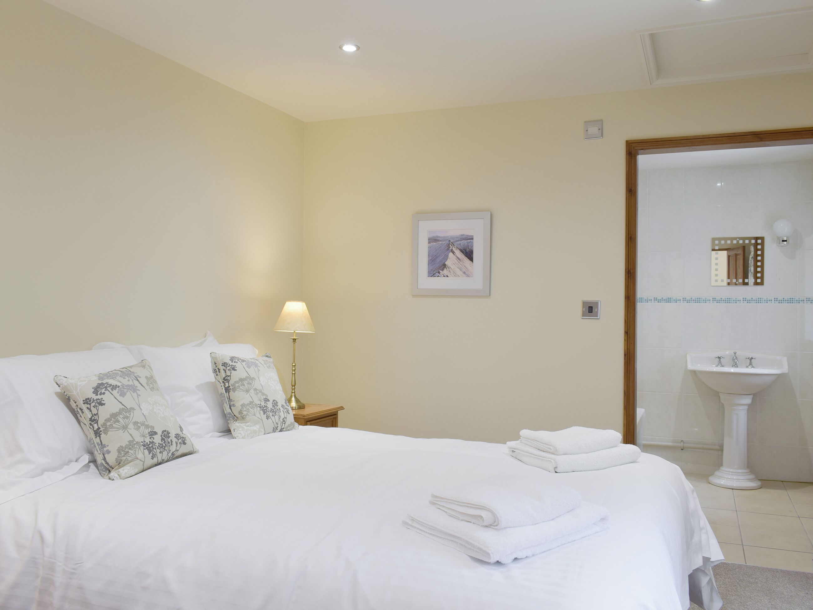 A neatly made bed with white linens and floral-patterned pillows, a bedside lamp, a framed artwork on the wall, and a bathroom with a sink and mirror.