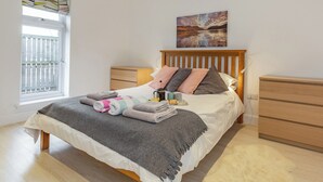 Een netjes opgemaakt slaapkamer met een bed, nachtkastjes, een raam met jaloezieën en een schilderij aan de muur.
