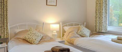 A twin room with two beds, a bedside table with a lamp, a framed picture on the wall, and a window with curtains.