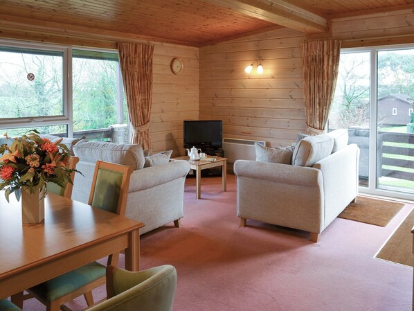 Ein gemütliches Blockhaus-Interieur mit zwei Sofas, einem Esstisch, einem Fernseher und großen Fenstern mit Vorhängen.
