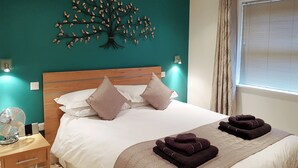 A neatly made bed with white linens and two pillows, a wooden nightstand with a fan, a teal wall with a tree decoration, and a window with blinds. - 7 bedroom accommodation in North Duffield, near York (North Duffield, near York)
