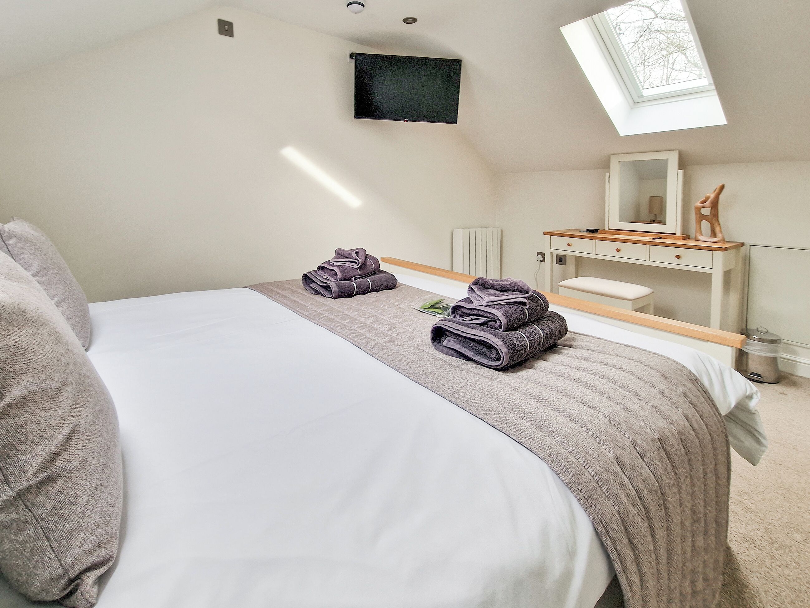A bedroom with a bed, two rolled towels, a bedside table, a mirror, and a skylight.