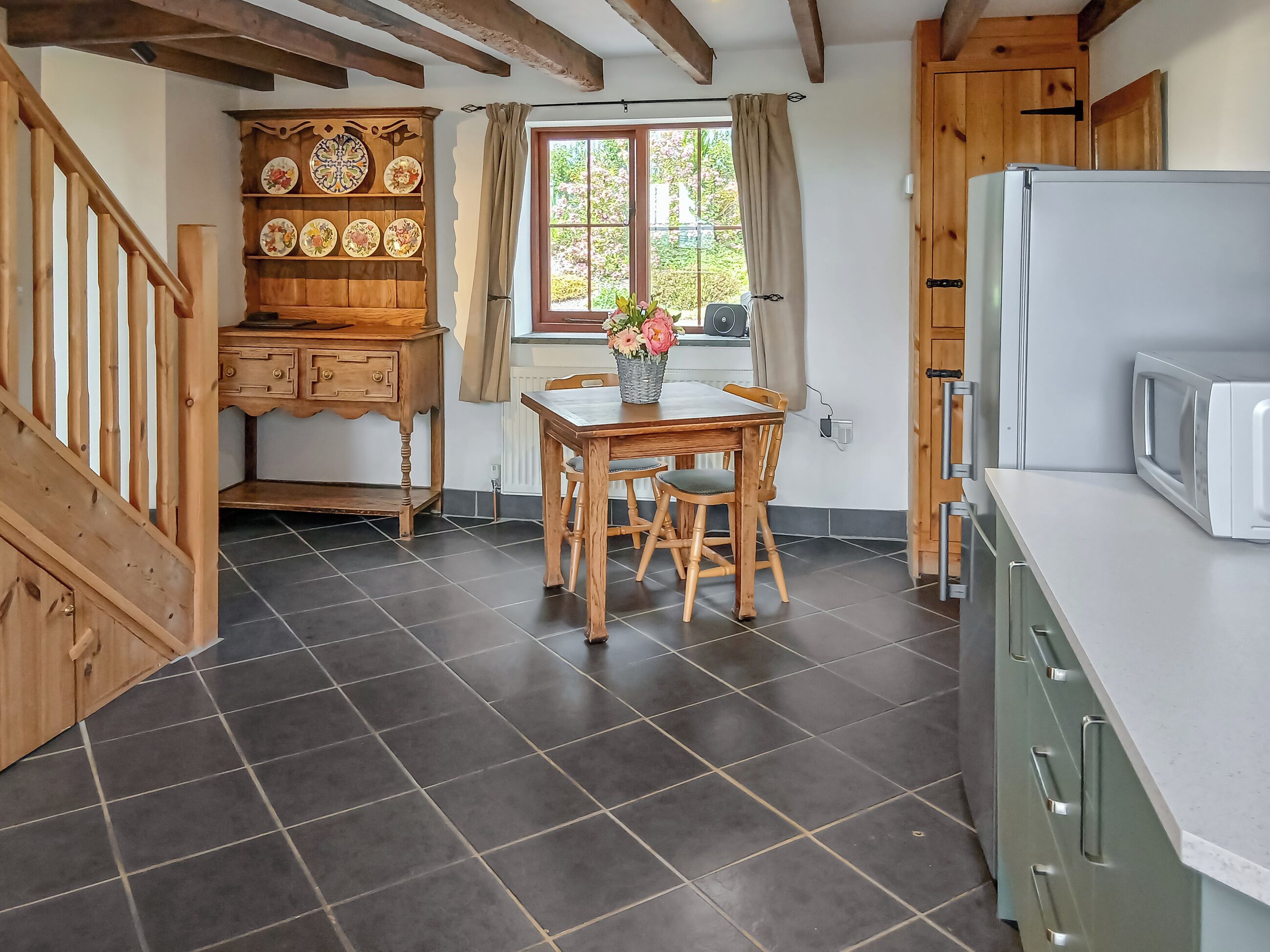 A kitchen with dark tiled flooring, wooden furniture, a dining table with chairs, a refrigerator, and a window with curtains.
