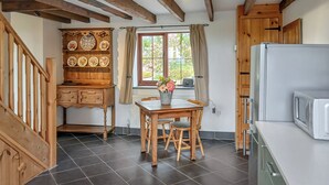 Una cocina con piso de baldosas oscuras, muebles de madera, una mesa de comedor con sillas, un refrigerador y una ventana con cortinas.