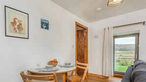 Un'area pranzo con un tavolo rotondo, sedie di legno e una ciotola di frutta. Si vede la campagna attraverso una finestra.