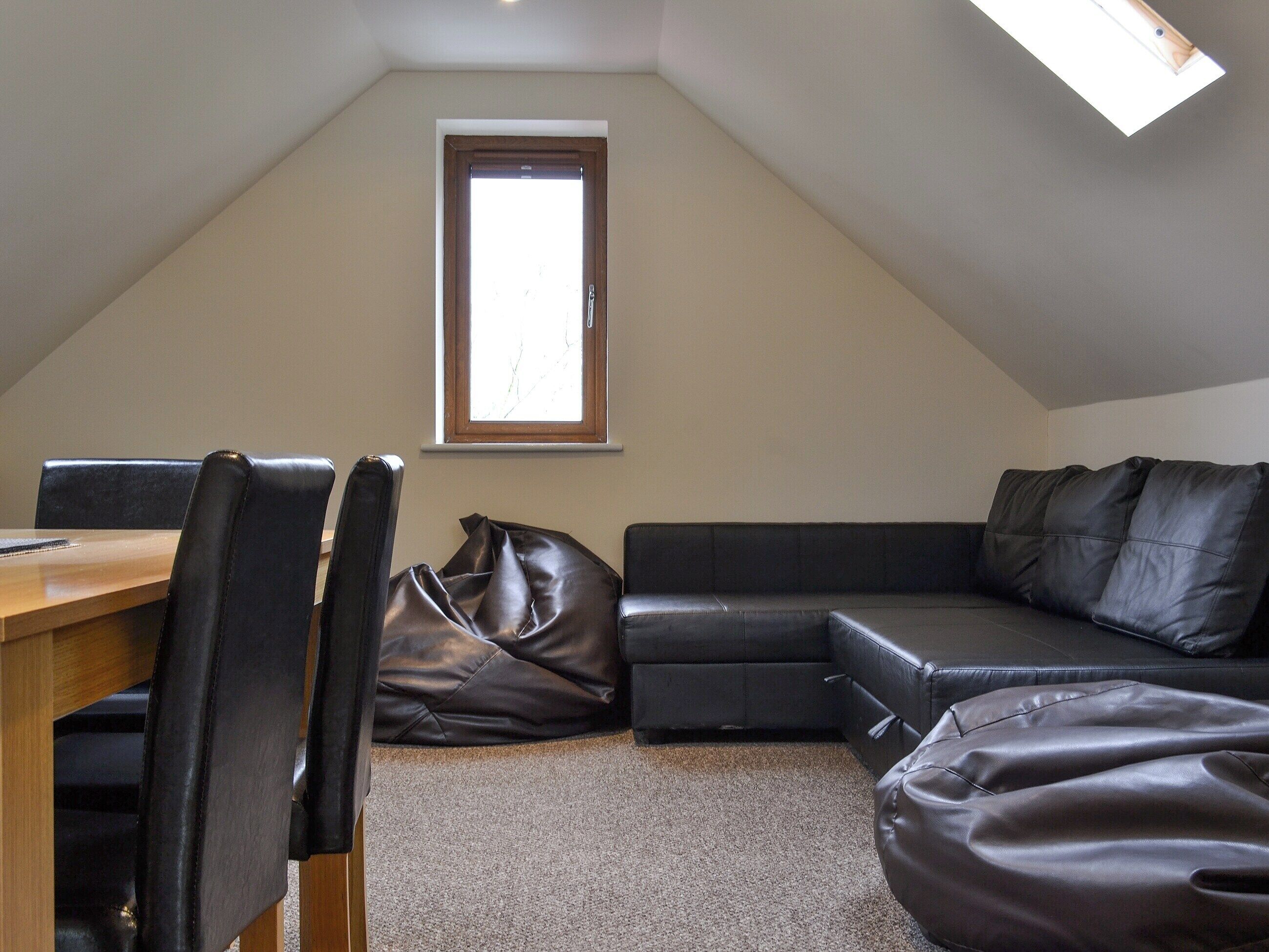 A room with a sloped ceiling, a window, a wooden table, a black leather couch, and a black leather bean bag.