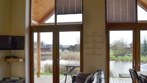 A living room with a leather sofa, a flat-screen TV, and a large window overlooking a pond and outdoor seating area. - 2 bedroom accommodation in Thorpe on the Hill, near Lincoln (Thorpe on the Hill, near Lincoln)