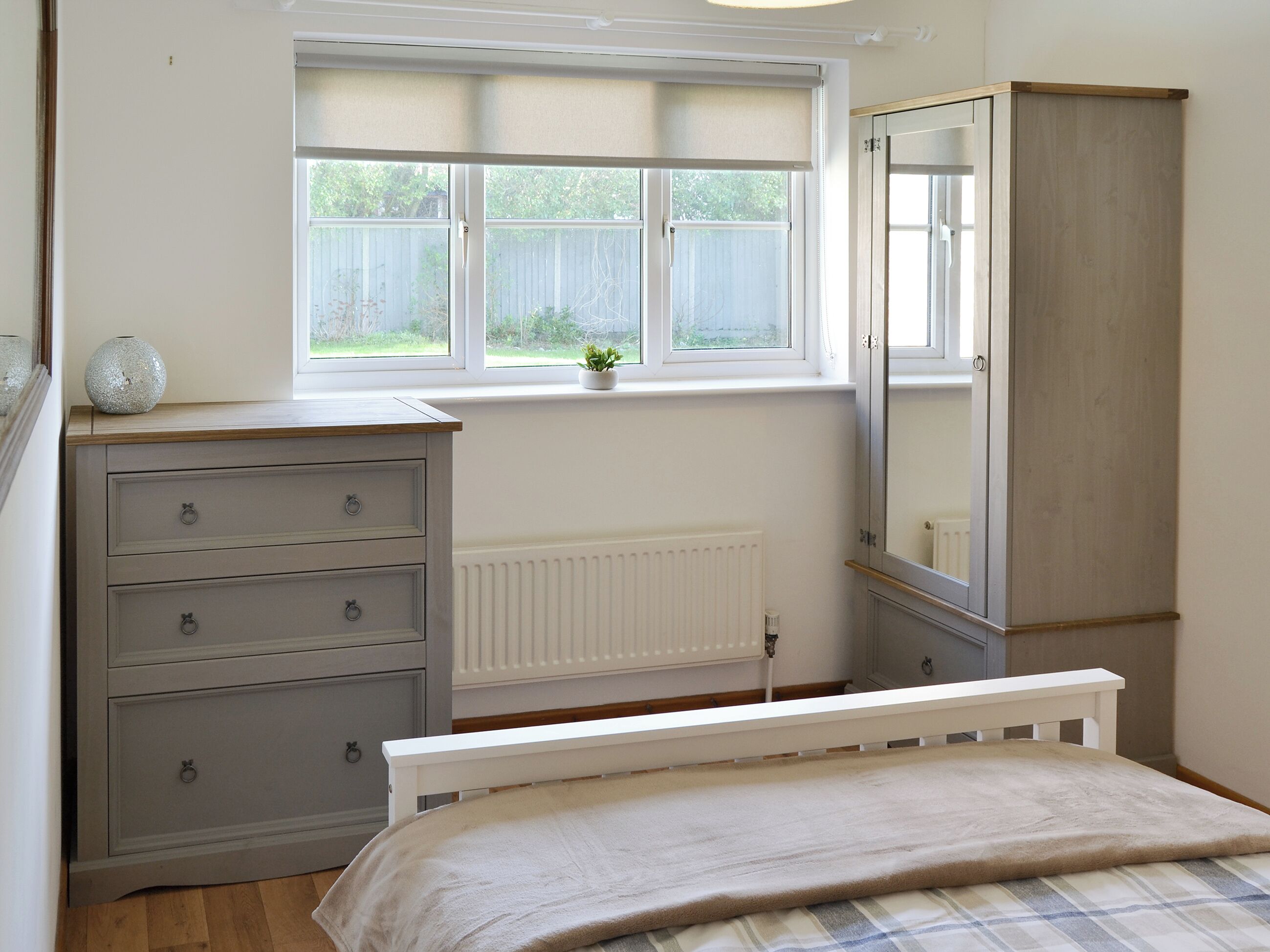 A bedroom with a bed, a dresser, a wardrobe, a window with a view of the outside, and a radiator under the window.