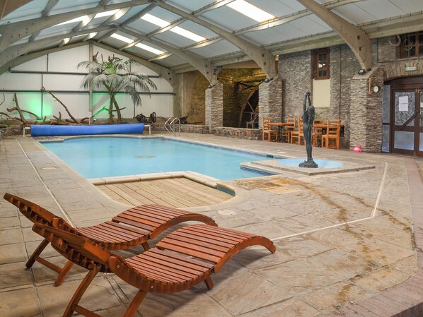 An indoor swimming pool with wooden loungers, a stone wall, and a palm tree.