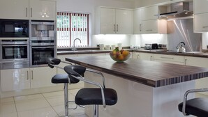 A modern kitchen with stainless steel appliances, a central island with bar stools, and a window with vertical blinds. - 6 bedroom accommodation in Stepaside, near Saundersfoot (Stepaside, near Saundersfoot)