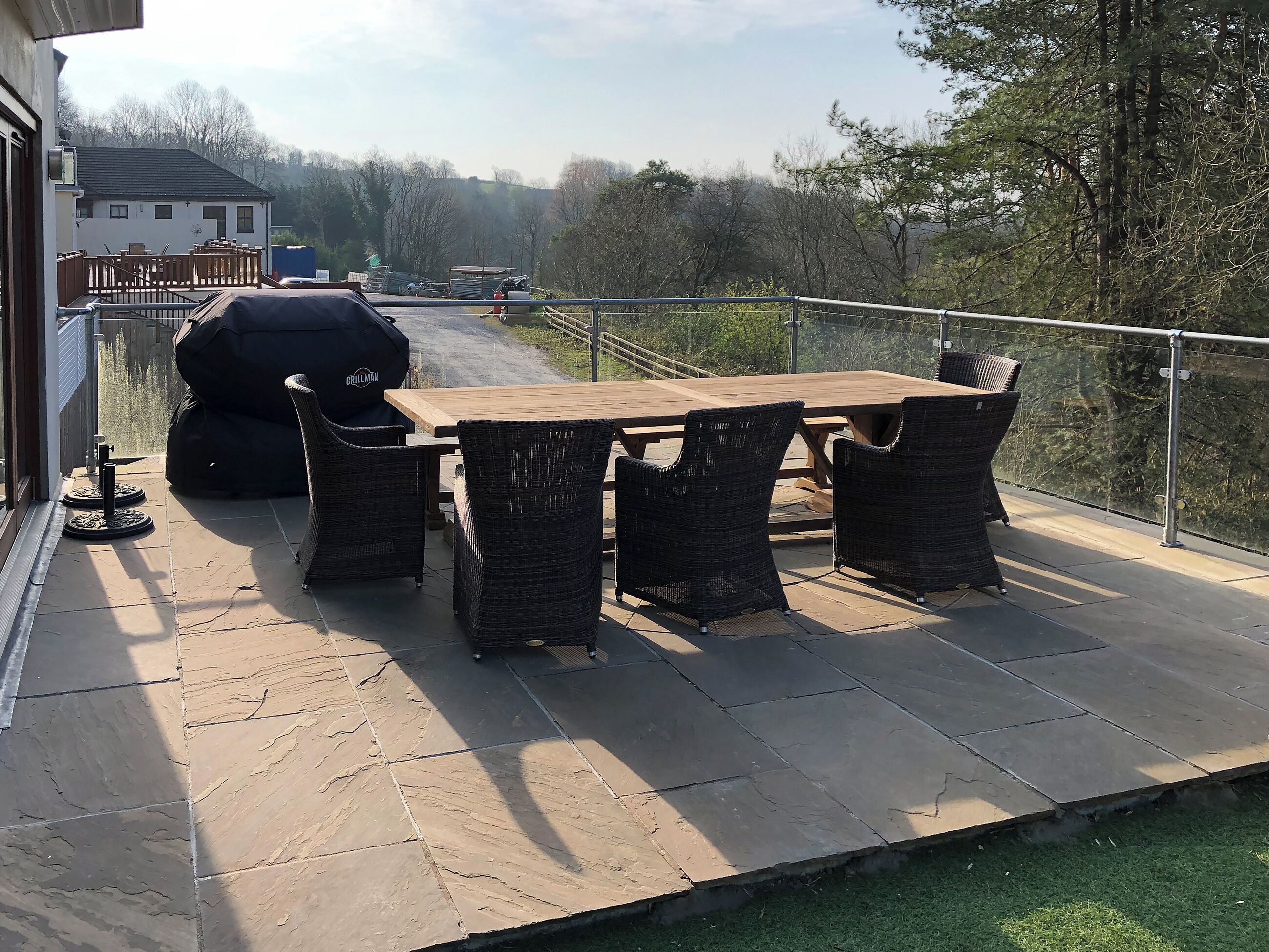 An outdoor patio with a wooden table and wicker chairs, a grill, and a view of a house and trees.