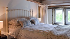Una camera da letto con soffitto in legno, un letto con lenzuola a motivi floreali, un comodino con una lampada e una finestra con tende.