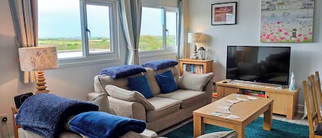 A living room with a sofa, a coffee table, a TV, and a view of the sea.