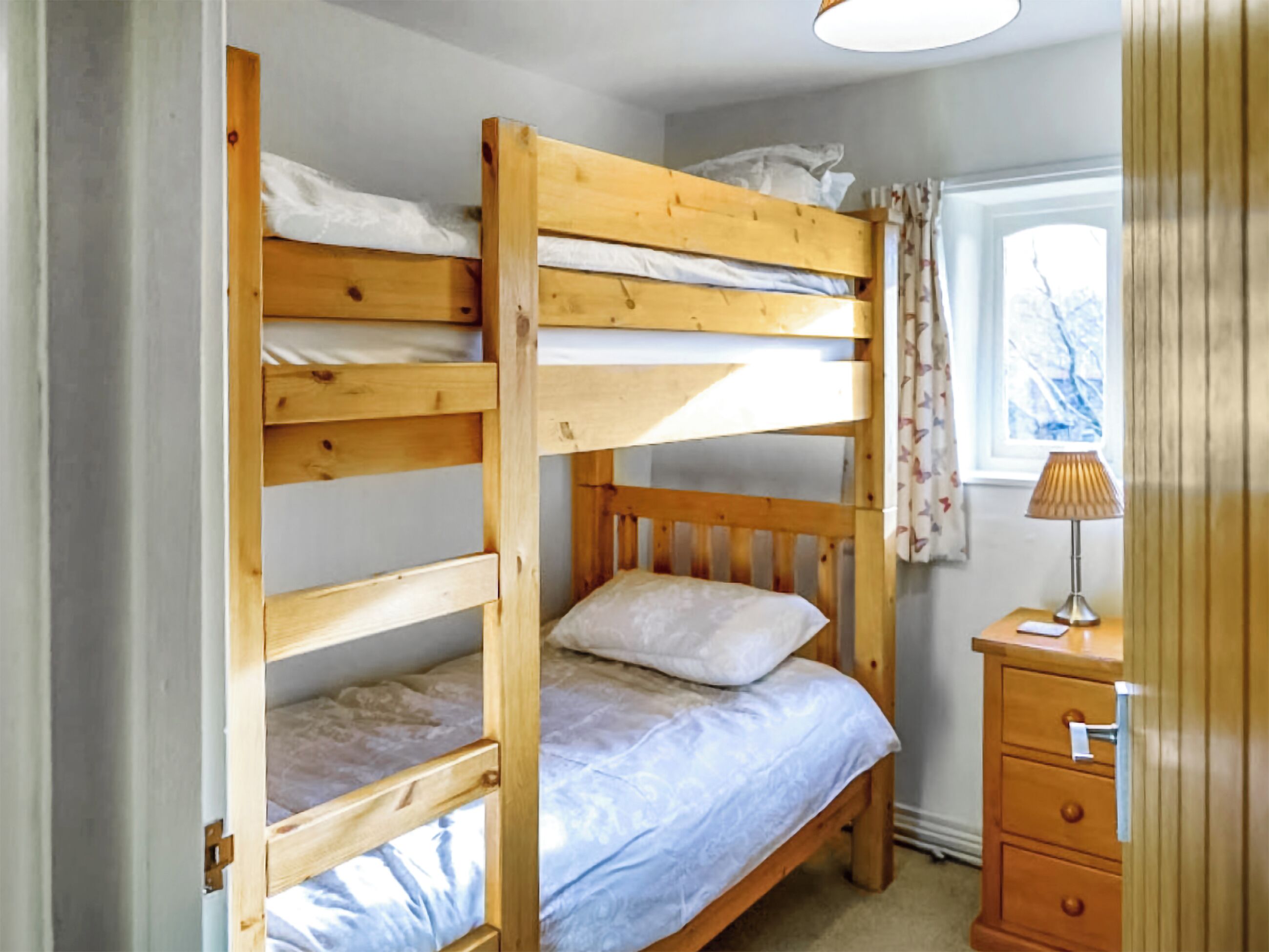 A bunk bed room with a wooden headboard, a bedside table with a lamp, and a window with curtains.