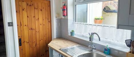 A compact kitchen with a wooden door, a washing machine, a sink with a window above, and a small window with a plant.