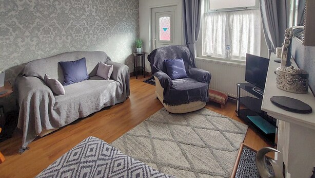 Una sala de estar con un sofá, un sillón y un televisor. Hay una ventana con cortinas, un radiador y una pequeña mesa con una planta.
