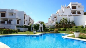 A swimming pool surrounded by white buildings with balconies, a clear sky, and well-maintained greenery. - Golf Hills Marbella - Beautiful decorated incl. lounge terrace (Estepona)