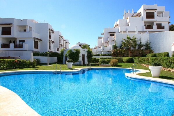 Une piscine entourée de bâtiments blancs dotés de balcons, un ciel dégagé et une végétation bien entretenue.