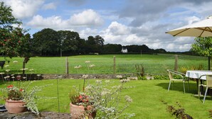 A fenced outdoor area with a grassy field, a white house in the background, and a table with an umbrella. - 2 bedroom accommodation in Dwyran, near Llanfairpwllgwyngyll (Dwyran, near Llanfairpwllgwyngyll)