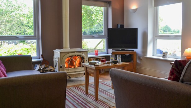 A cozy living room with a fireplace, a television, and large windows offering a view of greenery. - 2 bedroom accommodation in Dwyran, near Llanfairpwllgwyngyll (Dwyran, near Llanfairpwllgwyngyll)