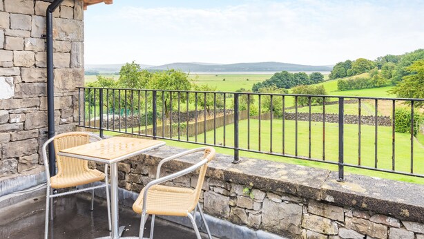 Ein Balkon mit Steinmauern, einem Tisch, zwei Stühlen und Blick auf eine grüne Landschaft mit Feldern und Bäumen.