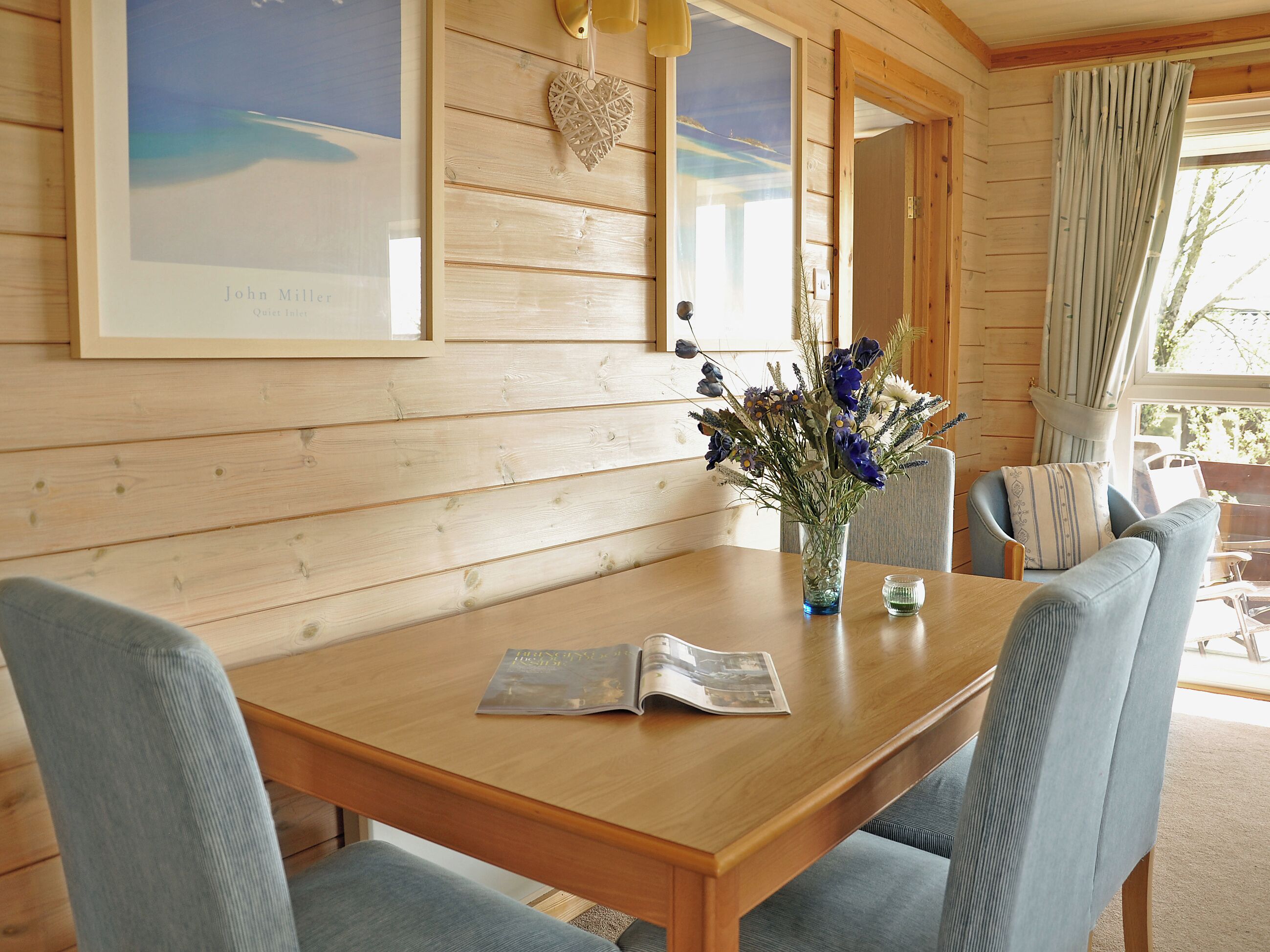 A wooden interior with a dining table, chairs, a vase with flowers, framed artwork, and a window with curtains.