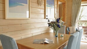 Un intérieur en bois comprenant une table à manger, des chaises, un vase avec des fleurs, des œuvres d’art encadrées et une fenêtre avec des rideaux.