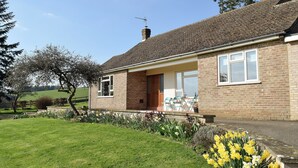 A single-story brick house with a tiled roof, a front porch with a table and chairs, and a garden with daffodils. - 3 bedroom accommodation in near Chipping Warden (near Chipping Warden)