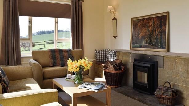 A cozy living room with a fireplace, a sofa, a coffee table with flowers, and a view of the outdoors. - 3 bedroom accommodation in near Chipping Warden (near Chipping Warden)