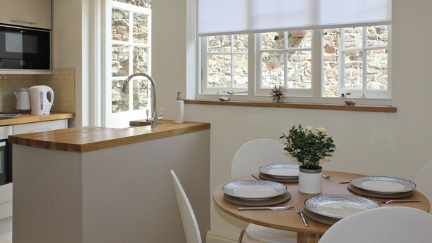 A kitchen with a built-in oven, a sink, and a window with a view of a stone wall. There is a dining area with a table set for four, plates, and a potted plant. - A great base for seaside enthusiasts. (Deal)