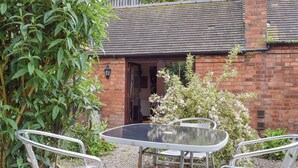 An outdoor patio with a glass table and metal chairs, surrounded by greenery and a brick building with a wooden door. - 2 bedroom accommodation in Upper Welland, near Malvern (Upper Welland, near Malvern)