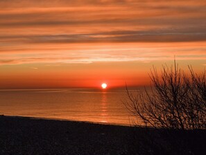 Un coucher de soleil sur une mer calme, avec en silhouette, une plage et une végétation clairsemée au premier plan.