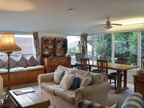 A cozy living room with a sofa, a wooden coffee table, a dining table with chairs, and a large window offering a view of greenery outside. - 4 bedroom accommodation in Pagham, near Chichester (Pagham, near Chichester)