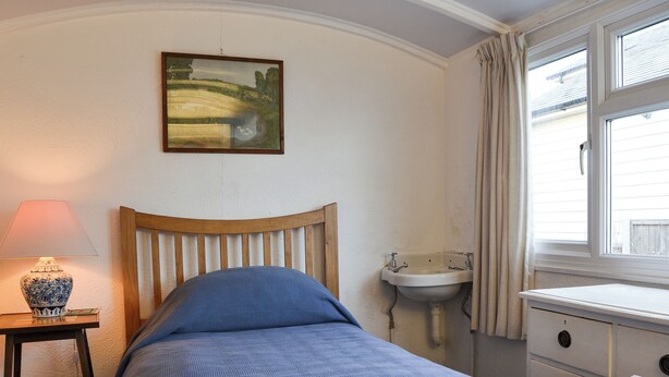 A bedroom with a wooden bed, a bedside table with a lamp, a wall-mounted painting, a window with curtains, and a small sink. - 4 bedroom accommodation in Pagham, near Chichester (Pagham, near Chichester)